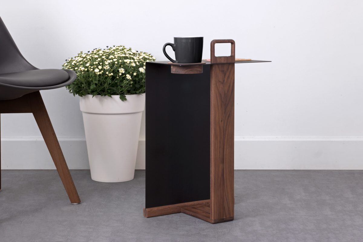 Side angle of walnut and black side table holding a black cup, near plant and chair